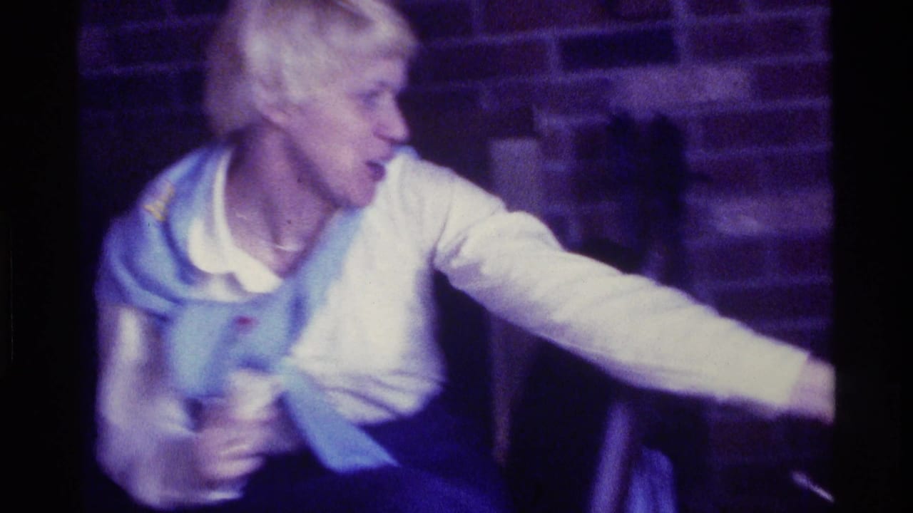 Blonde adult woman seated indoors and reaching to the side.