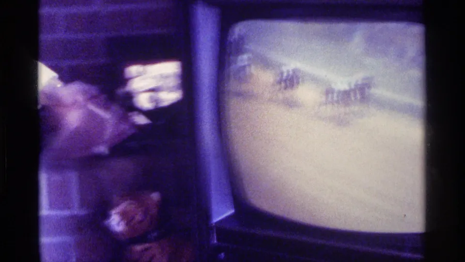 A television displays horses racing on a track while a blurred seated person appears beside the set in the Kentucky home interior.