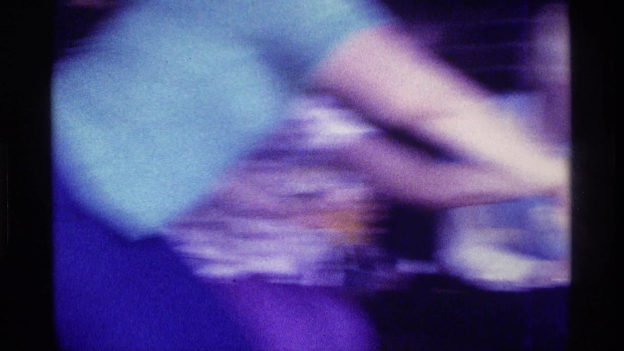 A heavily blurred 8mm frame shows close, fast movement among adults in the dim family-room setting, with clothing, arms, and a possible drinking glass visible.