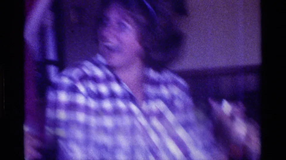 A woman in a plaid shirt appears to smile or laugh while holding a glass in the low-light home-movie interior associated with the supplied Kentucky 1976 clip metadata.