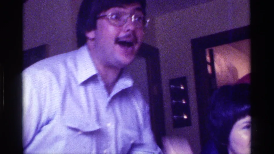 A man wearing glasses and a light shirt laughs broadly in a private Kentucky home interior, with another person partly visible at the lower right and a wall opening behind him.