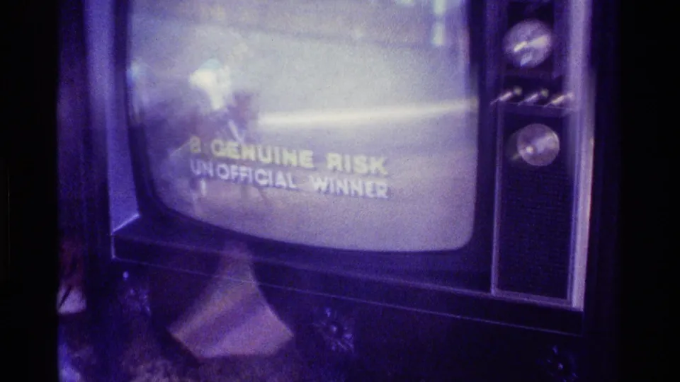 A filmed television screen shows race footage and a broadcast graphic reading 8-GENUINE RISK UNOFFICIAL WINNER.