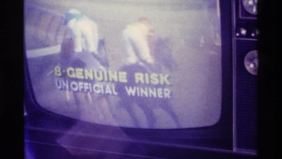 Close filmed-off-screen frame of riders on a television broadcast with the visible text 8-GENUINE RISK UNOFFICIAL WINNER.