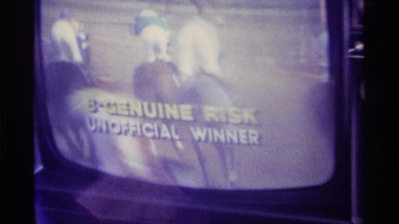 Television screen showing a horse race with text reading Genuine Risk unofficial winner.