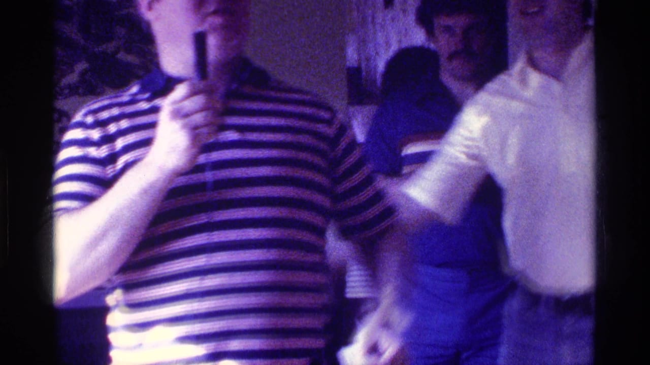 In 1976 Kentucky home-movie footage, a man in a striped polo holds a microphone during an indoor party with other adults nearby.