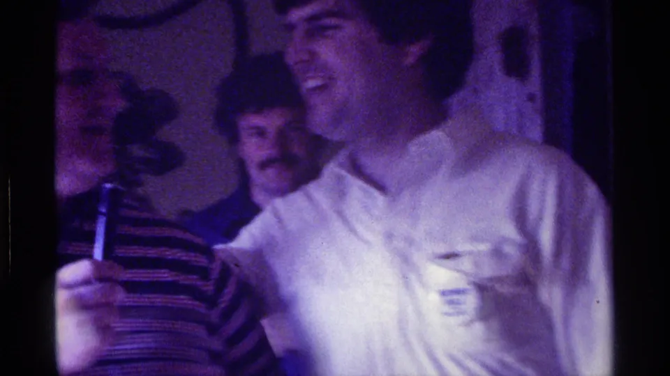 A laughing man in a white shirt stands close to the microphone while guests in the background watch inside a private Kentucky party setting.