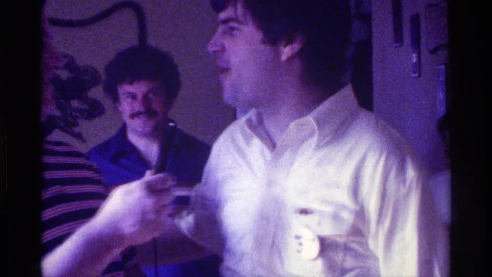 A smiling man in a white shirt with a campaign-style button faces a microphone during a 1976 Kentucky indoor party, with another guest behind him.