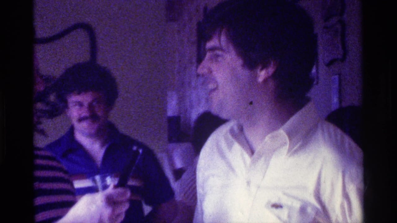 Man in a white shirt laughs while another man in a striped shirt stands behind him.