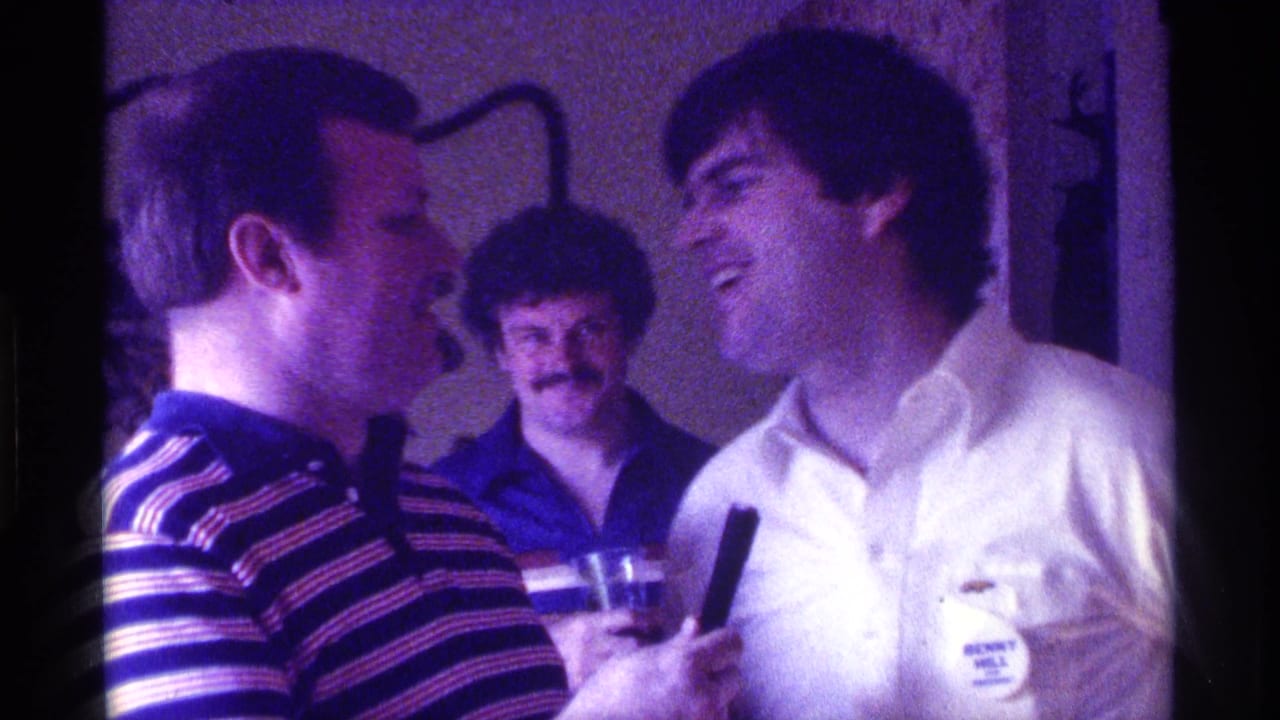 Man in a striped polo holds a microphone toward a laughing man in a white shirt.