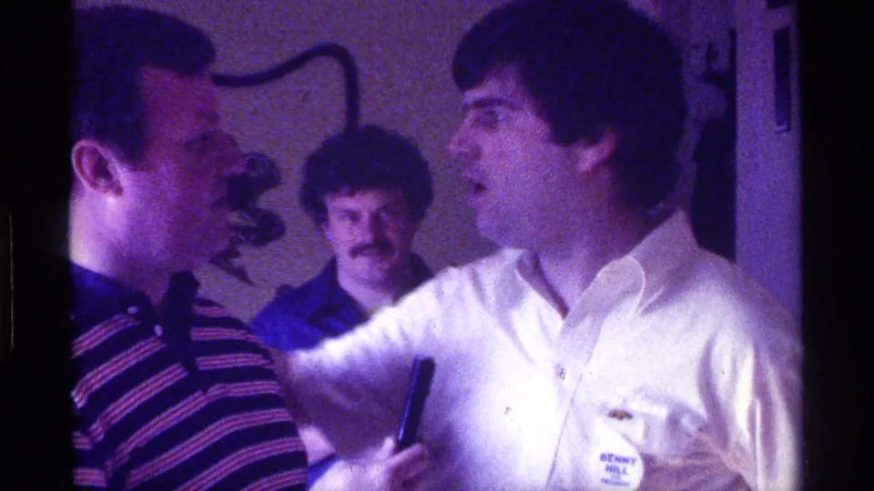 Two adult men face each other in close conversation, with a microphone between them and another man watching in the background.