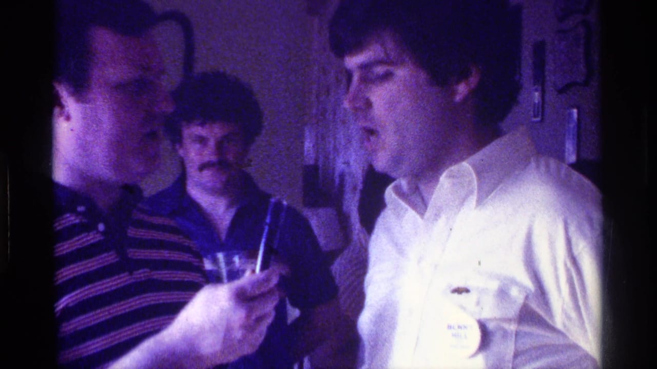 Man in white shirt speaks toward a handheld microphone while two men stand nearby indoors.