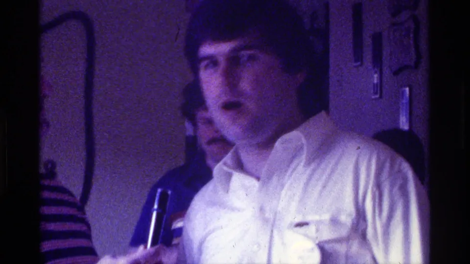 Closer 1976 Kentucky frame of the speaker in a white shirt, with a microphone at lower left and another man partly visible behind him.