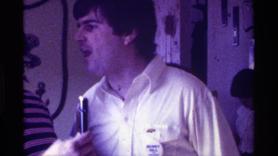 The speaker in a white shirt appears to smile or talk while a handheld microphone is raised near him during a 1976 Kentucky indoor gathering.