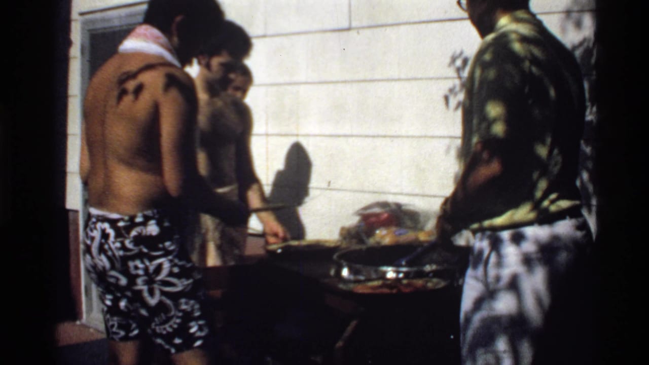 Three young men stand near a backyard grill beside a white-paneled house.