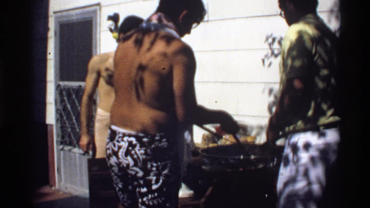 1978 California home-movie frame with a shirtless young man cooking at a backyard grill as two others stand near the house.
