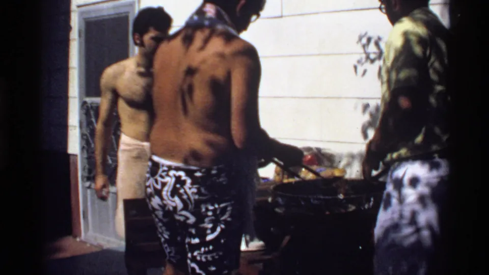 1978 California 8mm still of young men around a backyard barbecue, with house siding and a screen door in the background.