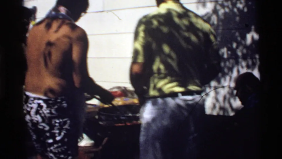 1978 California backyard barbecue frame with two young men at the grill and a partly visible seated figure at the right edge.