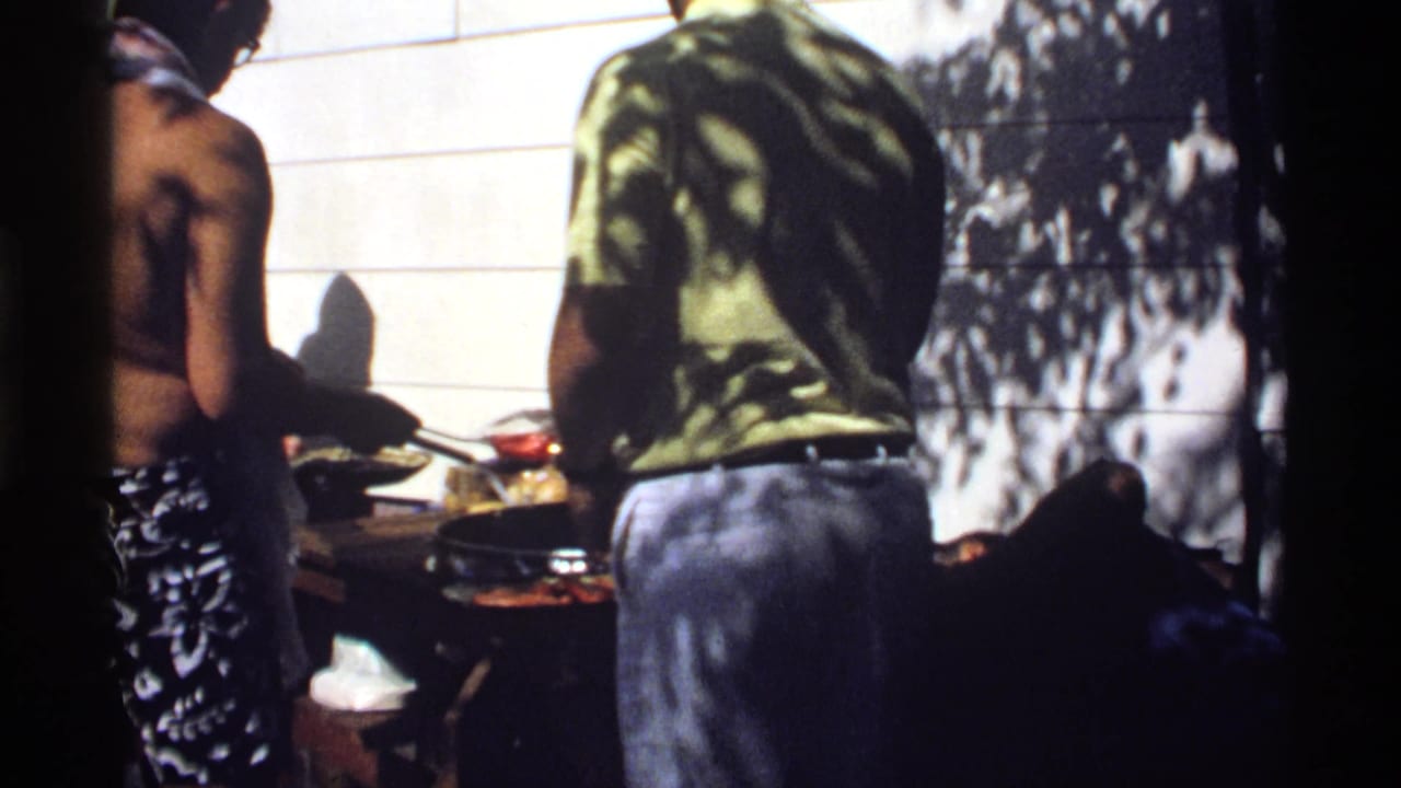 1978 California 8mm frame of shirtless men in swimwear standing near a backyard grill in dappled sunlight.