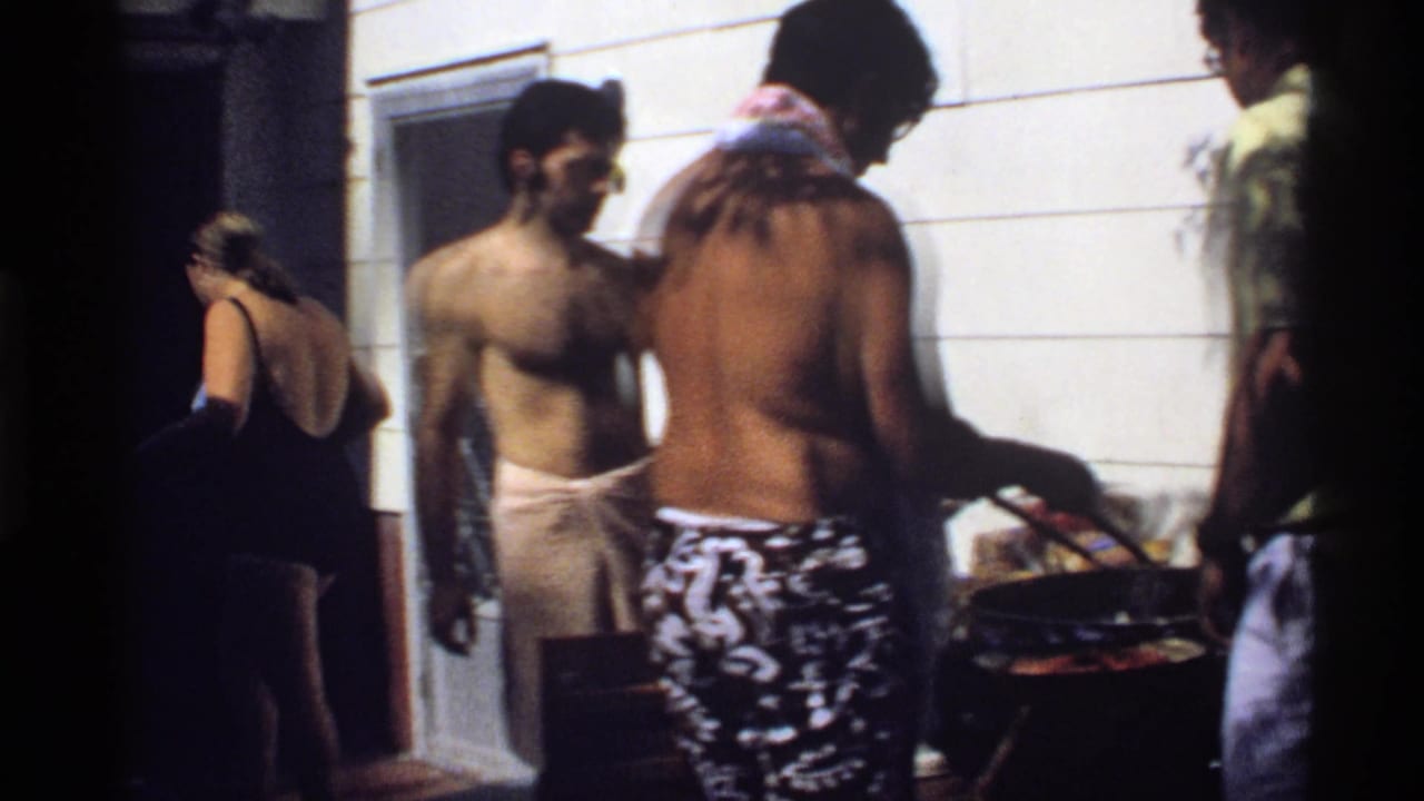 A woman in a dark swimsuit passes through the frame while men in swimwear stand at a California backyard cookout.