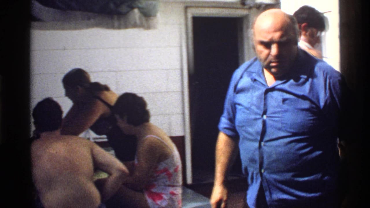 1978 California home-movie frame of adult men gathered outside by a house and grill during a backyard barbecue.