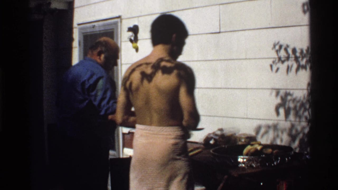 A shirtless man in a towel stands at a grill while another man in blue works nearby.
