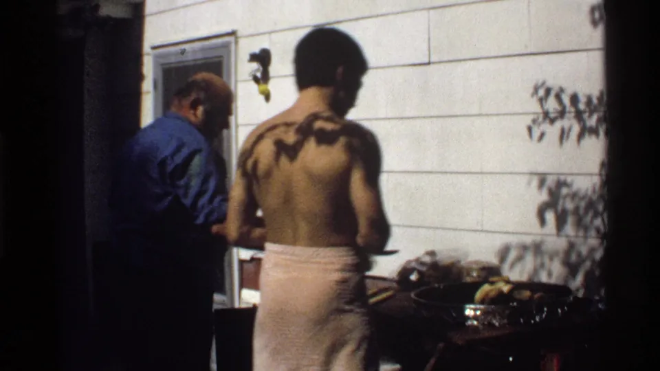 Adult men tend food at an outdoor grill beside a white-sided house in 1978 California home-movie footage.