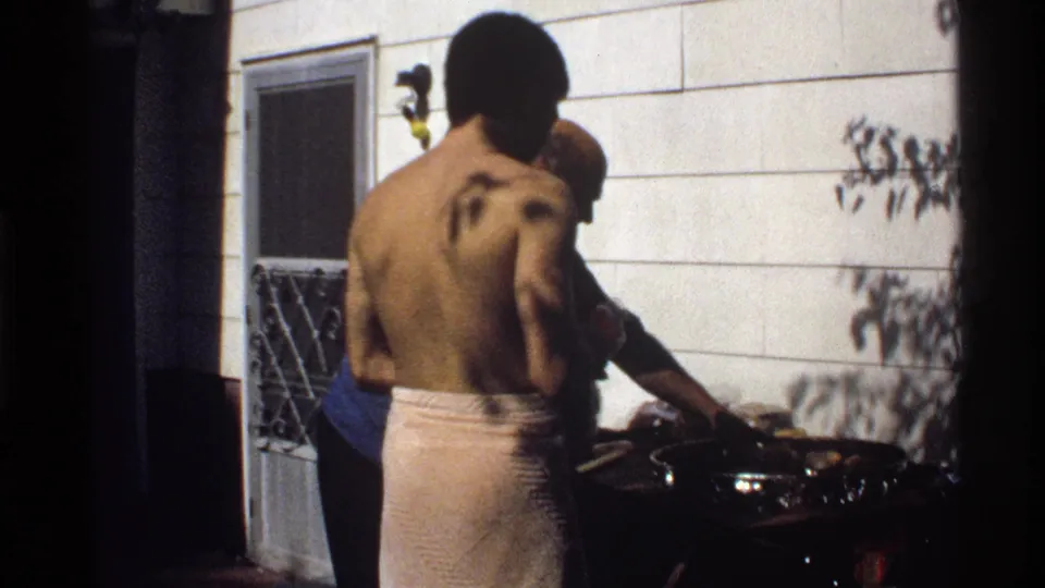 1978 California backyard barbecue frame with a shirtless adult man at the grill and another man partly visible nearby.