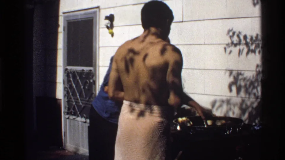In this 1978 California home-movie frame, a shirtless man tends food on a barbecue beside a sunlit house wall.