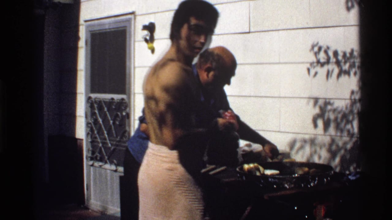 Shirtless man in a towel stands beside another man tending a backyard grill.
