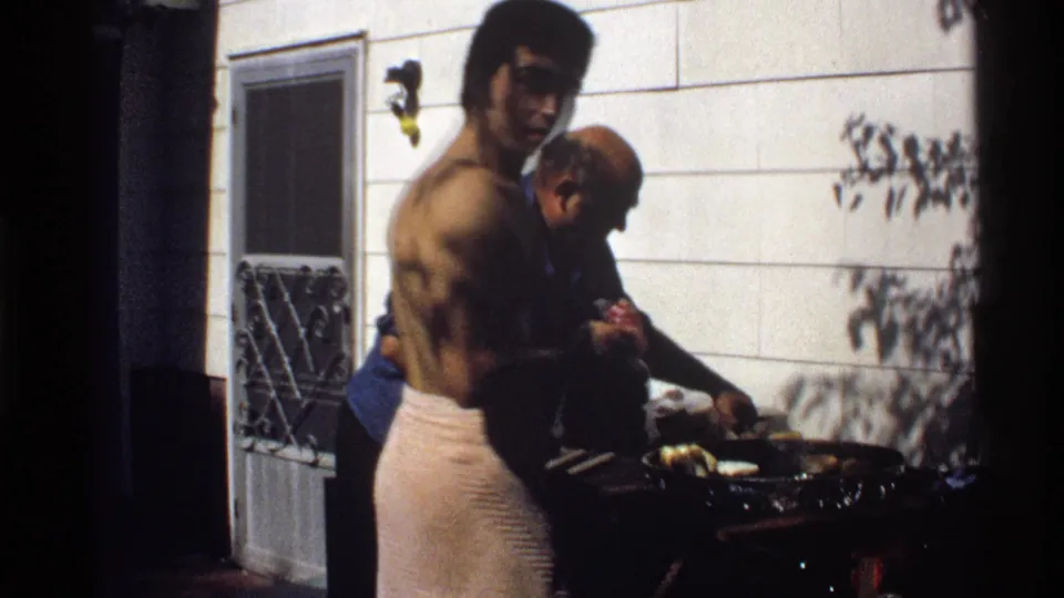 In 1978 California home-movie footage, a shirtless man in a towel stands near a man in blue as food cooks on an outdoor grill.