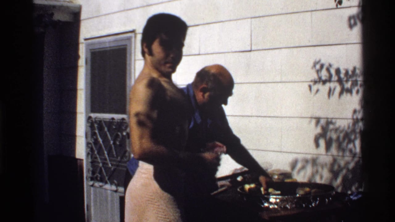 A shirtless man in a towel looks toward the camera as another adult man tends food on a backyard grill in California, 1978.
