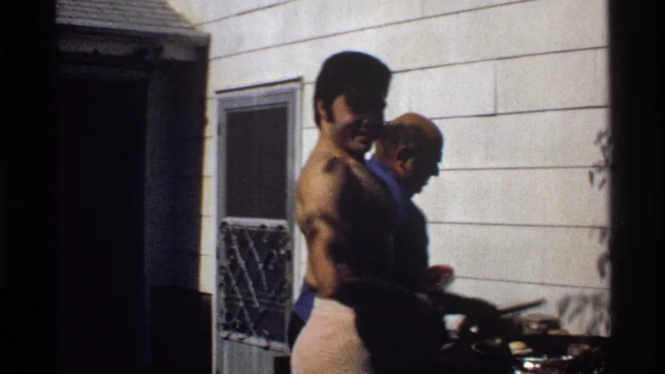 The backyard cooking scene continues, with the towel-wrapped man beside the grill and another man partly turned toward the food.