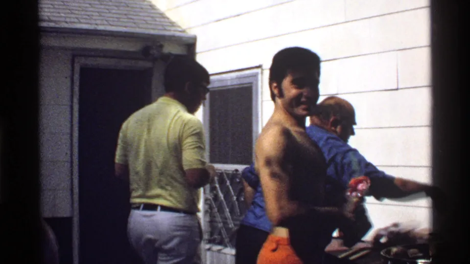 Three adult men gather around the grill, with a shirtless man holding an item and another man in a yellow shirt entering the scene.