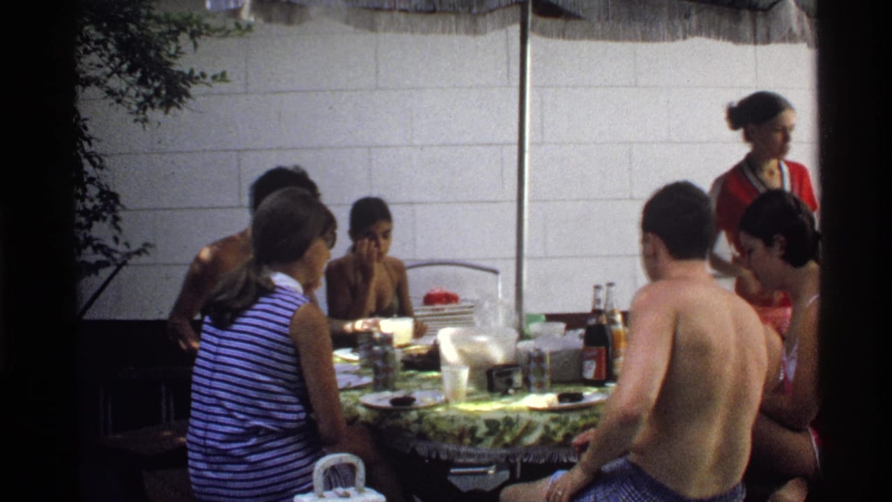 Young adults seated around an outdoor table with food and bottles near a light wall.