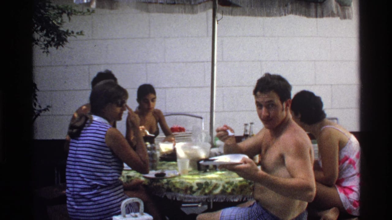1978 California home-movie frame with a shirtless man holding a plate beside seated companions at an outdoor table.