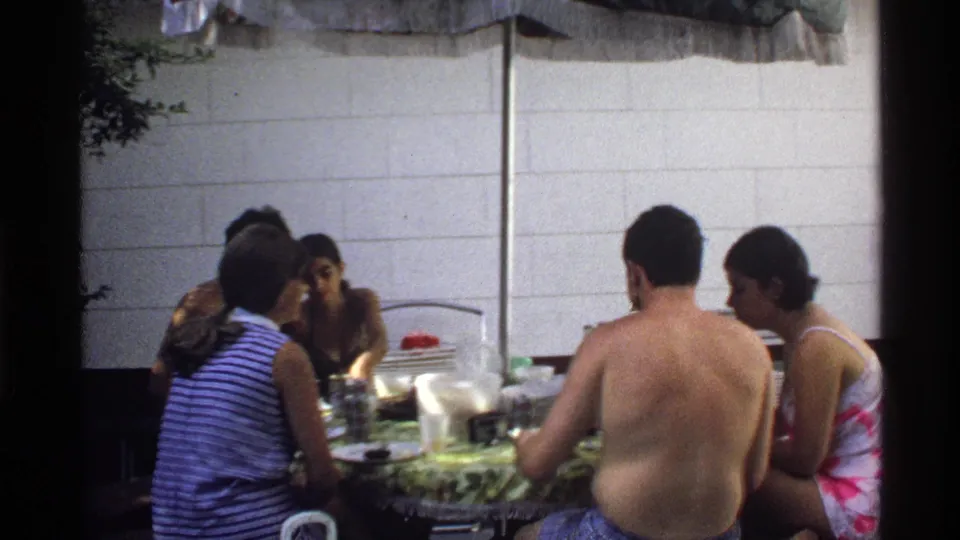 1978 California home-movie frame showing several young adults leaning in around a patio table during a casual meal.