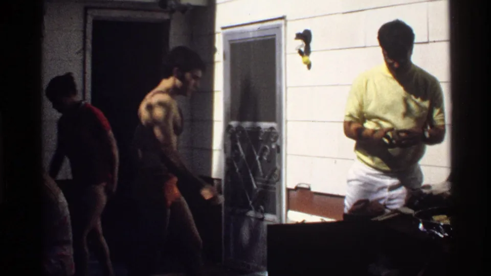 1978 California home-movie frame showing men near a doorway and exterior wall, possibly beside food preparation items.