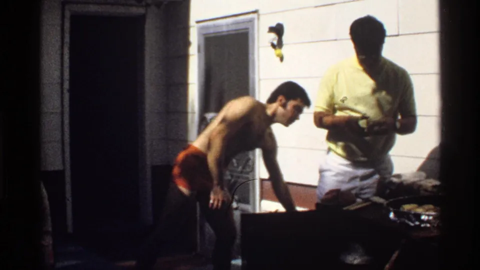 1978 California 8mm frame showing one shirtless man and one man in a yellow shirt preparing food at an outdoor grill.