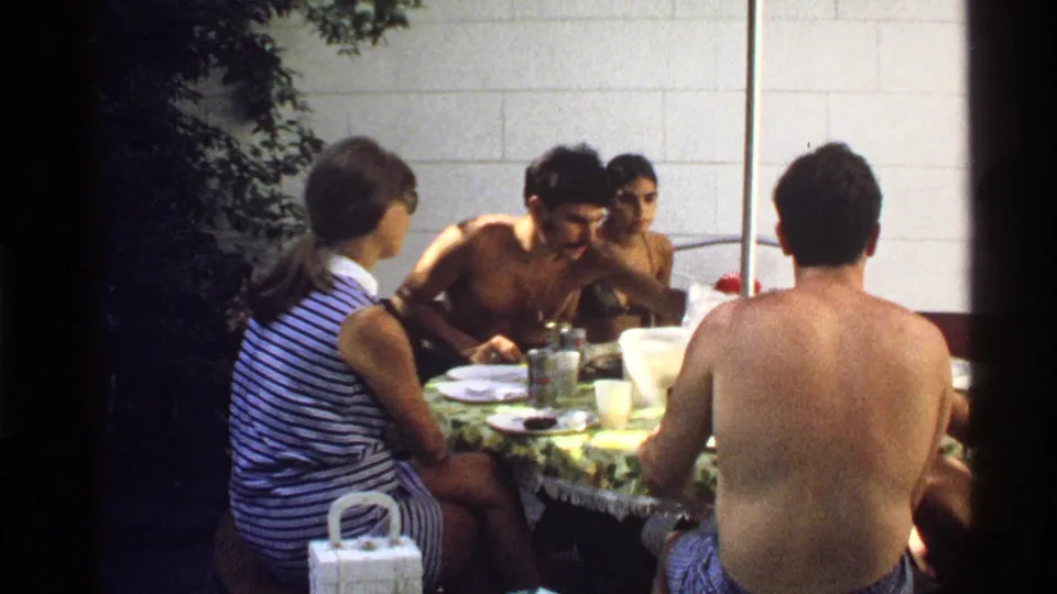 A 1978 California home-movie frame shows adults in swimwear and a striped top eating together at an outdoor table.