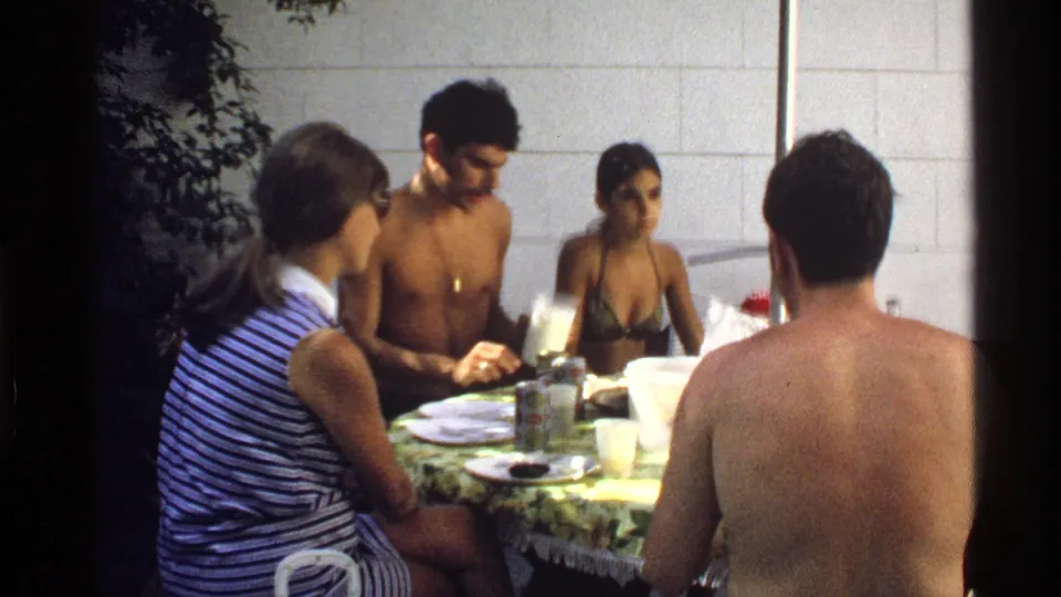 Adults sit closely around a backyard-style table in 1978 California, with swimwear, plates, pitchers, and drinks visible.