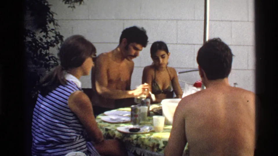 1978 California home-movie frame of young adults gathered at a backyard table with plates, cups, bottles, and a white block wall behind them.