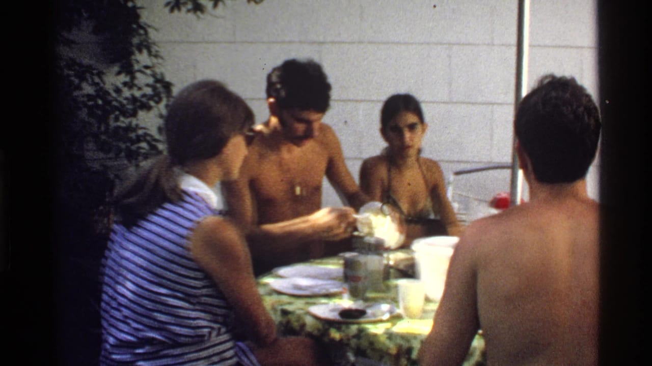 Young adults in a California backyard sit around a patterned table as a shirtless man handles food and others watch or converse.