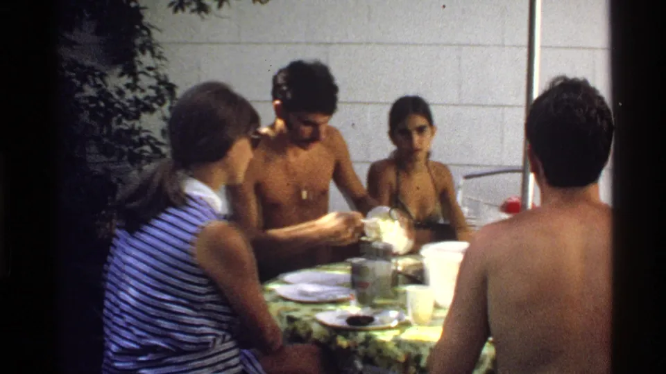 Young adults in a California backyard sit around a patterned table as a shirtless man handles food and others watch or converse.
