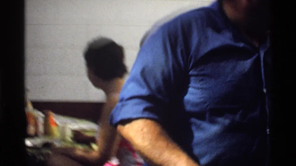A man in a blue shirt crosses close to the 8mm camera, partially obscuring a backyard table and seated figure in the background.