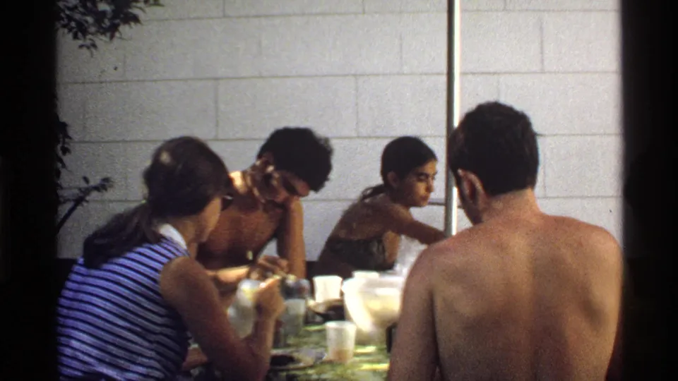 1978 California backyard picnic frame with young adults leaning around a table of cups, bottles, plates, and serving dishes in bright daylight.