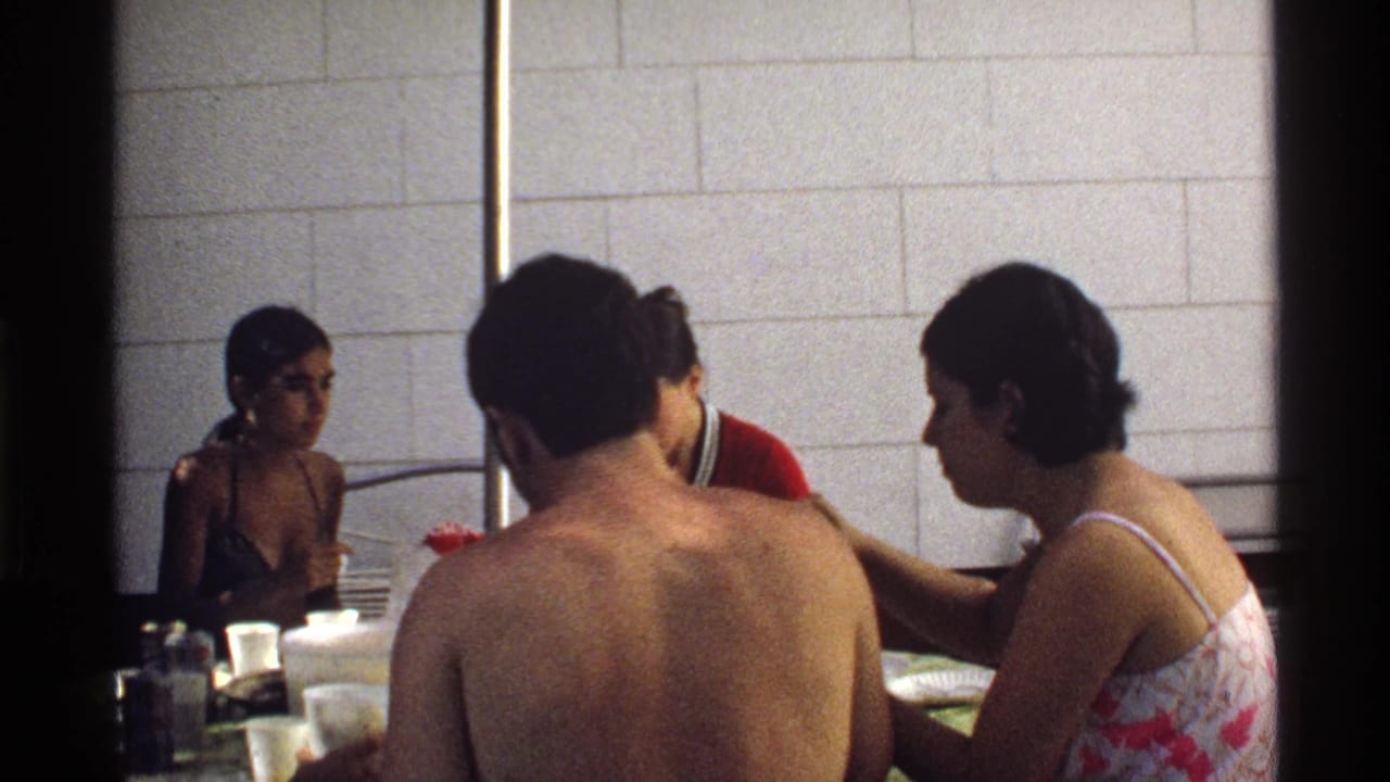 Adults seated around an outdoor table near a tiled wall, with a shirtless man seen from behind.
