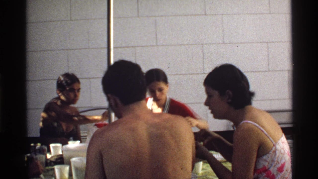 Adults in swimwear and casual summer clothes sit by a table in a California poolside setting, filmed on 8mm in 1978.