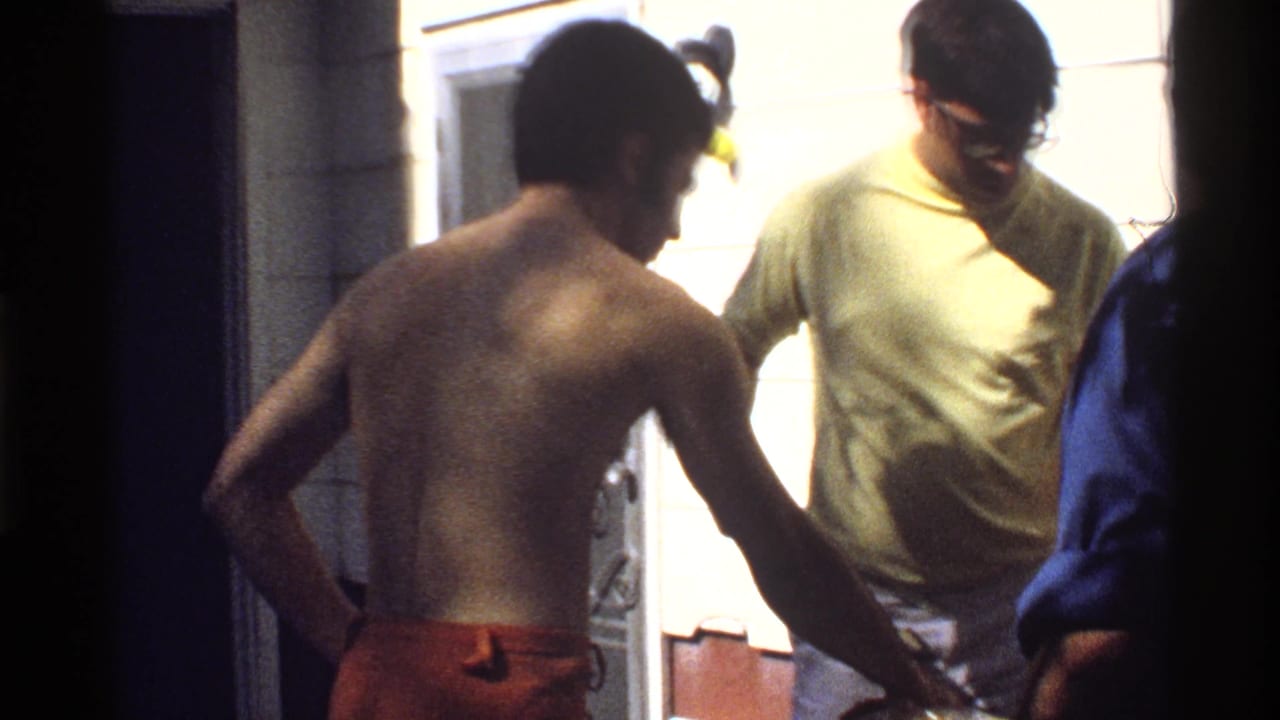 A shirtless man in orange swim trunks stands near a doorway with a casually dressed man, in 1978 California poolside home-movie footage.