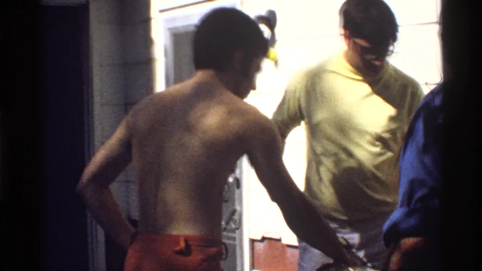 A shirtless man in orange swim trunks stands near a doorway with a casually dressed man, in 1978 California poolside home-movie footage.