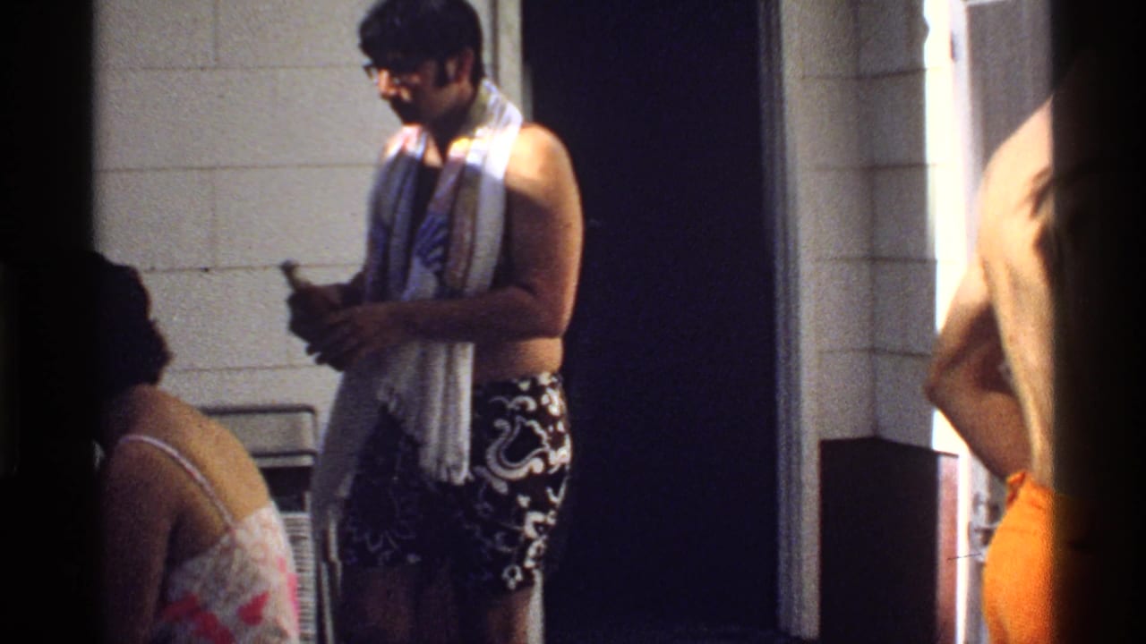 Man in patterned swim trunks stands near tiled wall while a seated woman in a floral swimsuit is partly visible.